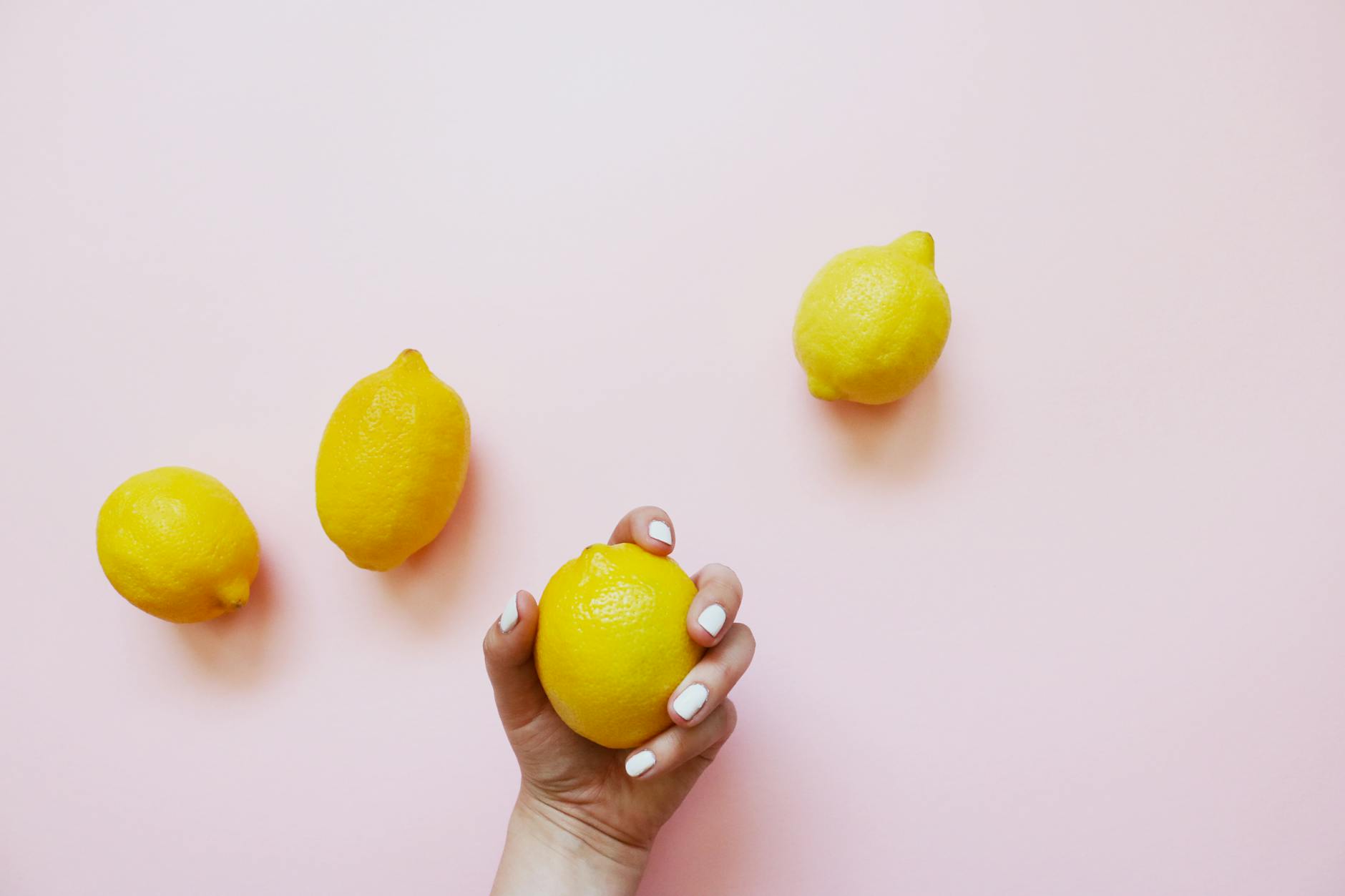 A hand holding a bright lemon on a soft pink background surrounded by additional lemons